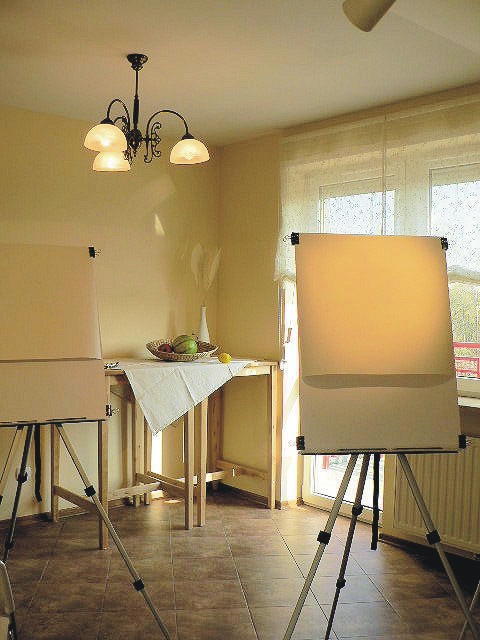 A bright art studio featuring two easels with blank canvases facing a window, a small table with art supplies, and a chandelier overhead.