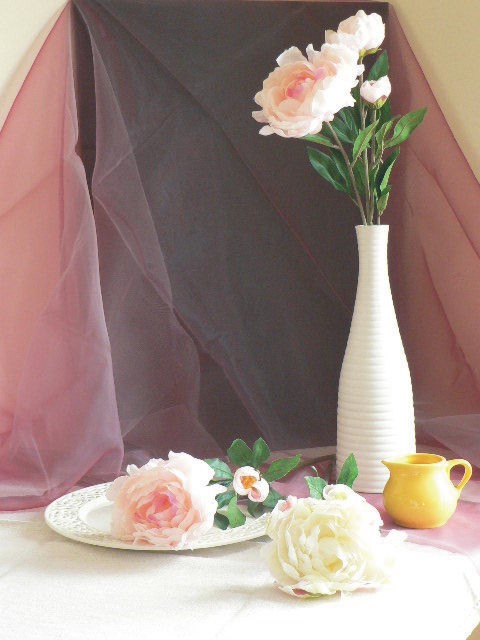 A still life composition featuring a white vase with pink flowers, a decorative plate with artificial blooms, and a yellow jug, set against a backdrop of layered fabrics in shades of pink and gray.