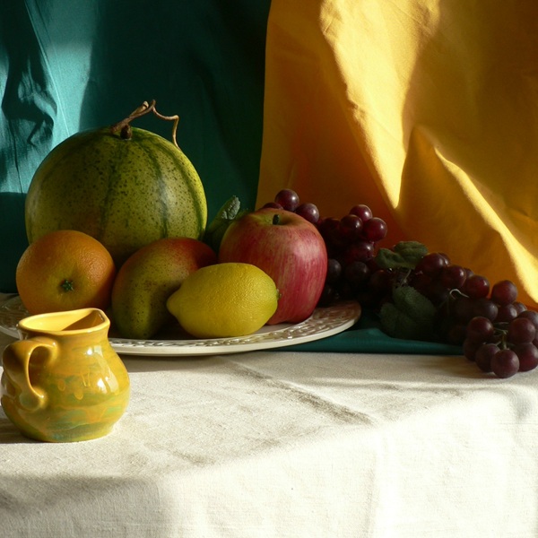 A still life arrangement featuring a green melon, orange, yellow lemon, red apple, and a cluster of grapes on a plate, with a small decorative jug in the foreground. The background consists of colorful fabric drapes in teal and yellow hues.