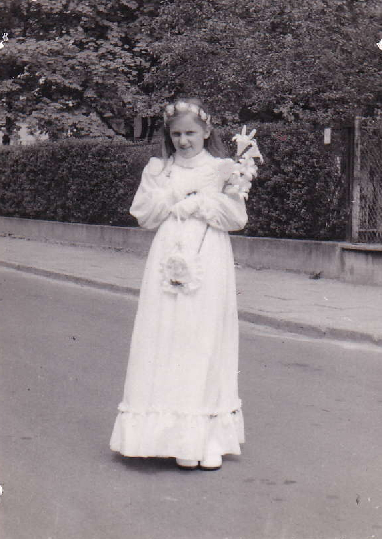 A young girl wearing a white dress and holding flowers, standing on a street with greenery in the background.