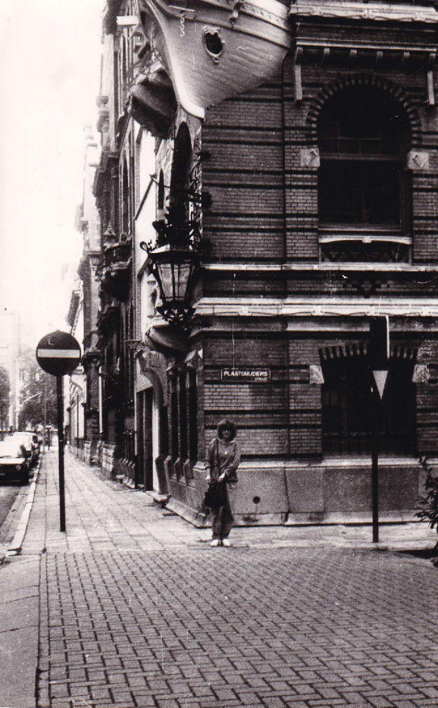 Black and white photograph of a woman standing on a cobbled street beside a historic brick building with decorative elements and a sign. A lamp post and parked cars are visible in the background.