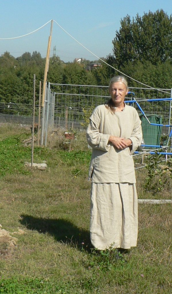 A woman standing in a grassy field, wearing a long light-colored dress and a matching top, with trees and a fence in the background.