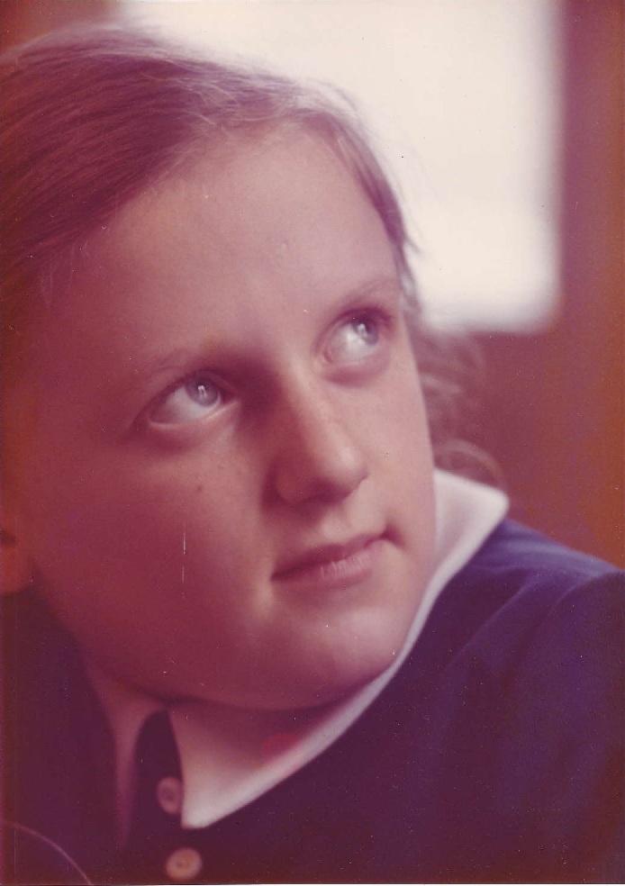 A close-up portrait of a young girl with blue eyes and light brown hair, looking slightly upwards. She is wearing a dark-colored shirt with a white collar.