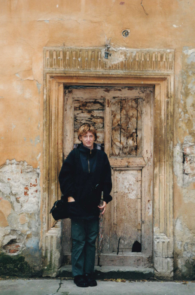 A person standing in front of an old, weathered doorway with intricate details, surrounded by peeling plaster and a faded wall.