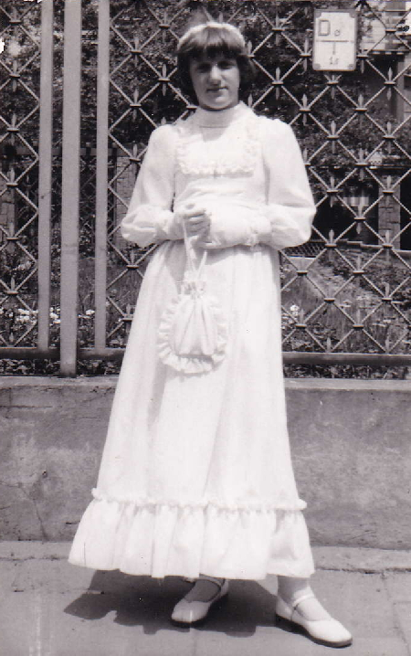 A black and white photograph of a young girl standing in front of a decorative metal fence, wearing a long white dress and holding a small bag.